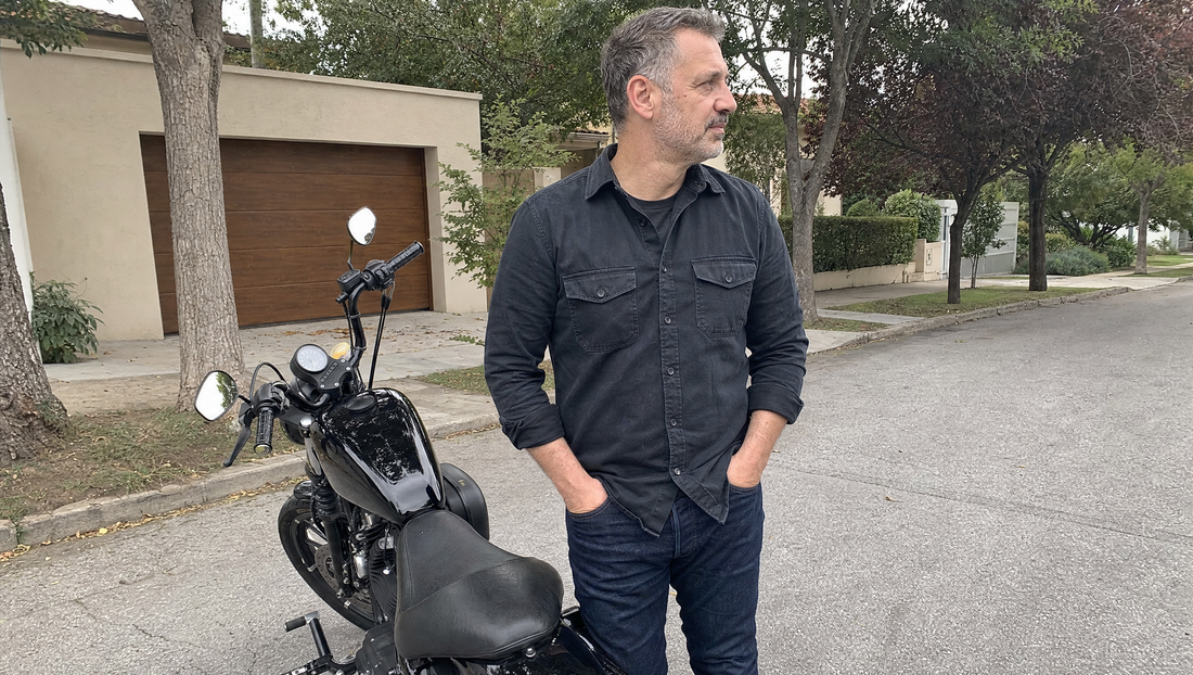 Motorcycle rider in dark casual armored streetwear standing beside a Harley cruiser on a quiet suburban street