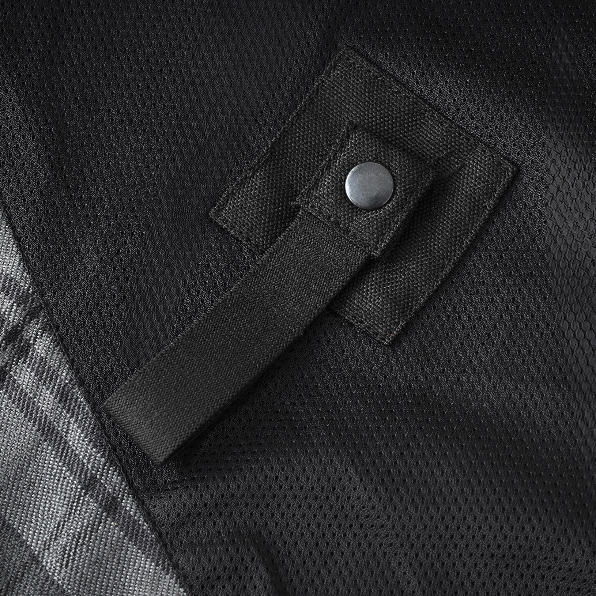 Close-up of a black mesh fabric surface with a black strap and metal snap fastener, plus a small portion of a gray plaid pattern at the lower left.
