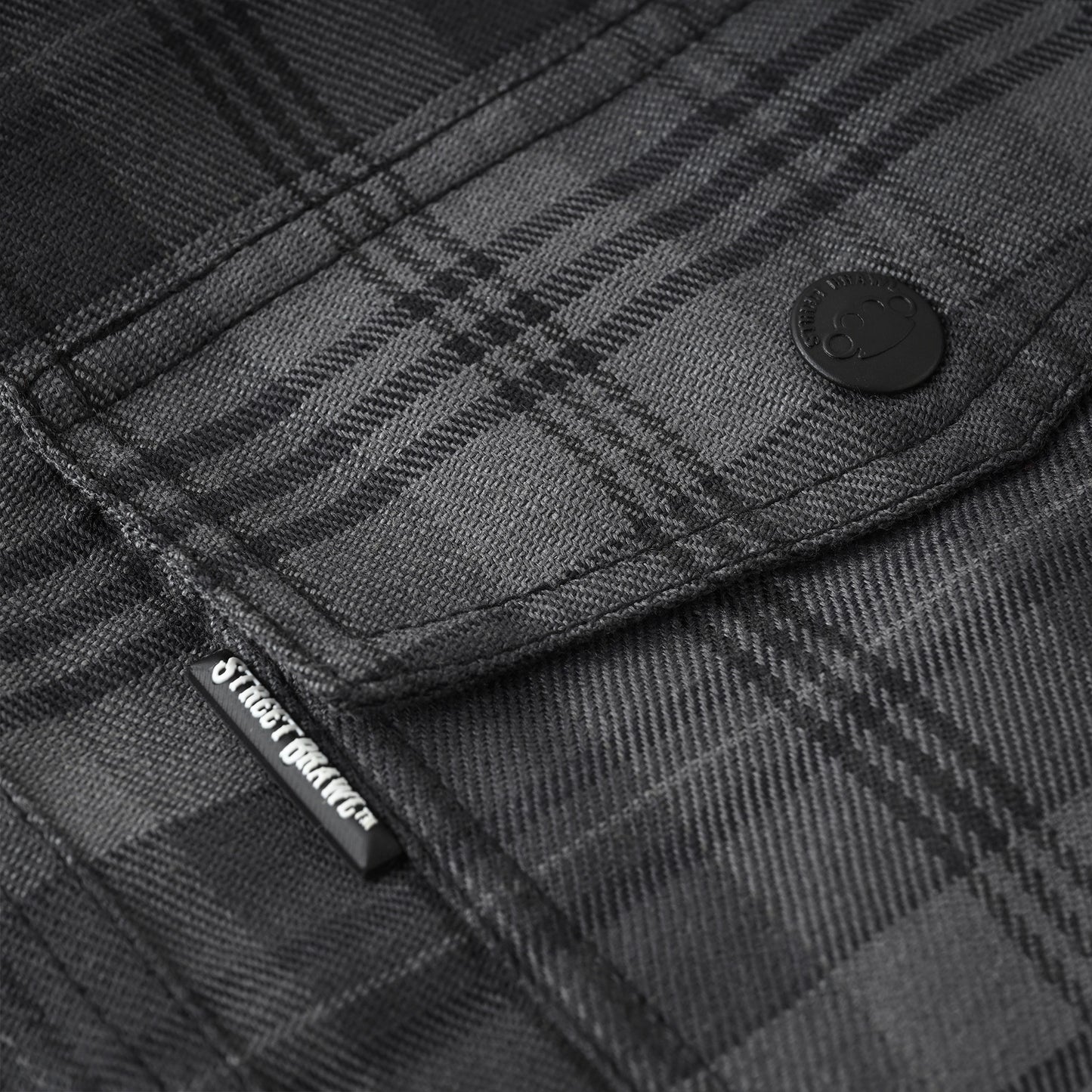 Close-up of a dark gray and black plaid flannel shirt fabric, showing a buttoned pocket with a black brand tab that reads Street Style.
