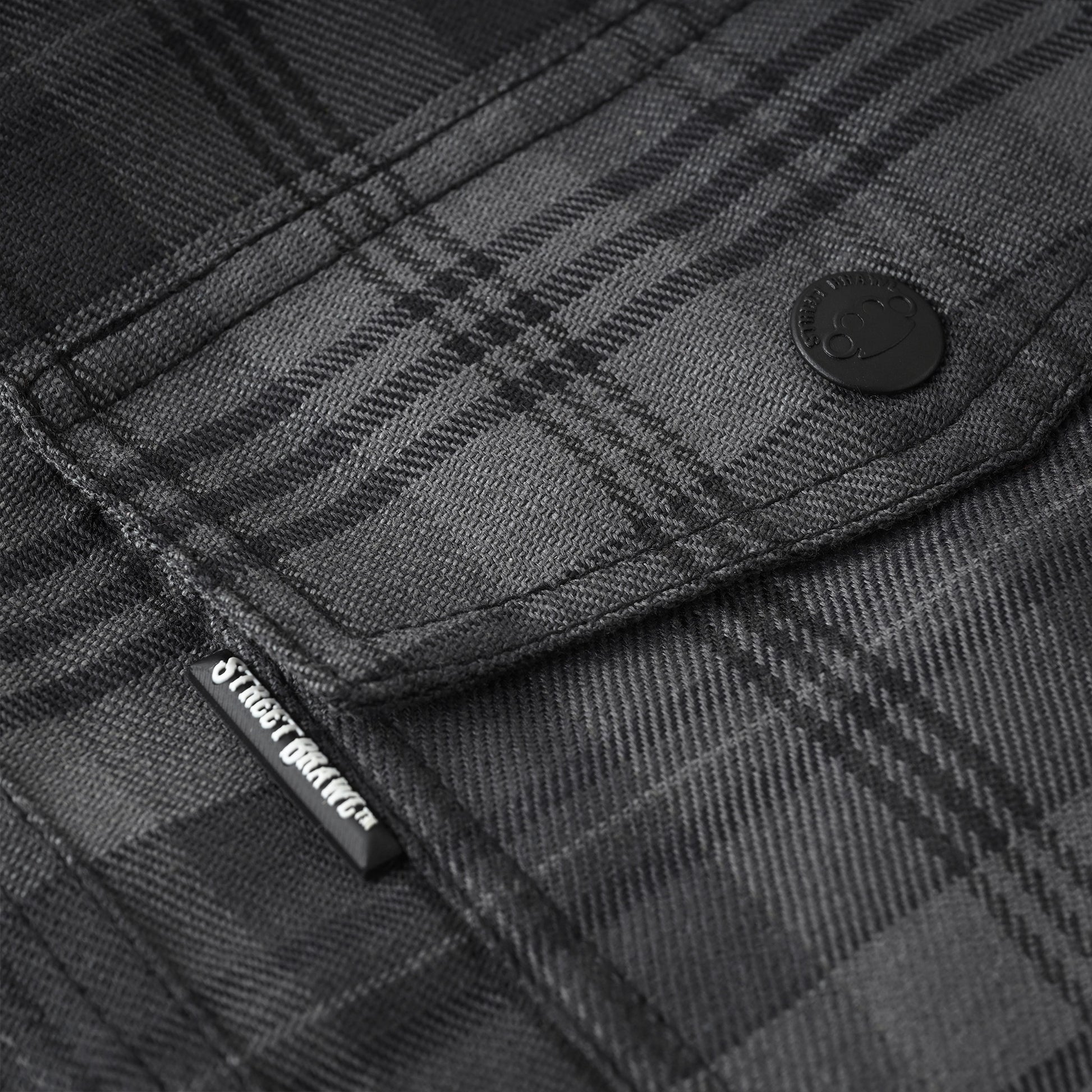 Close-up of a dark gray and black plaid flannel shirt fabric, showing a buttoned pocket with a black brand tab that reads Street Style.