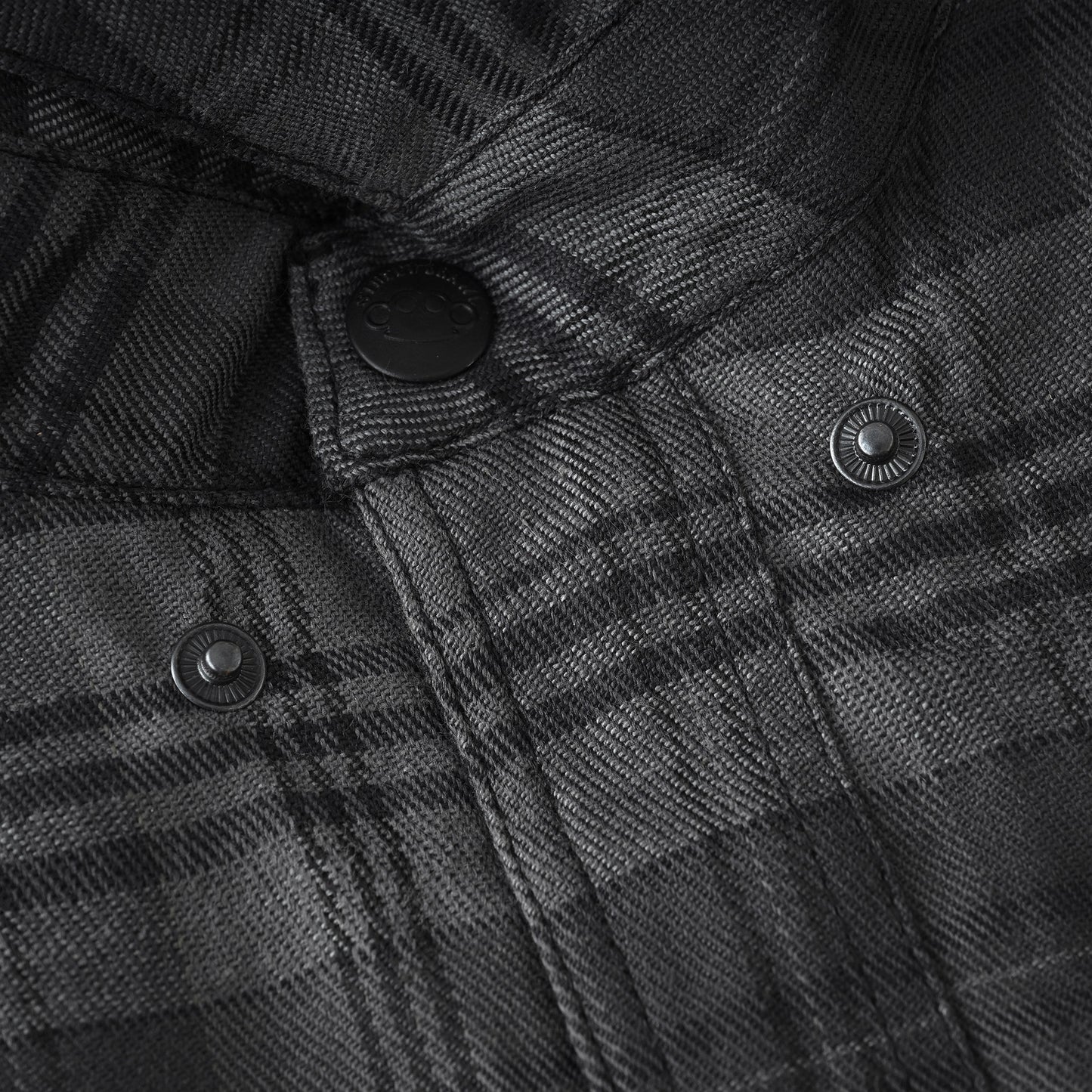 Close-up of a dark gray and black plaid fabric jacket or overshirt with black snap buttons, showing the detailed woven texture and stitching.