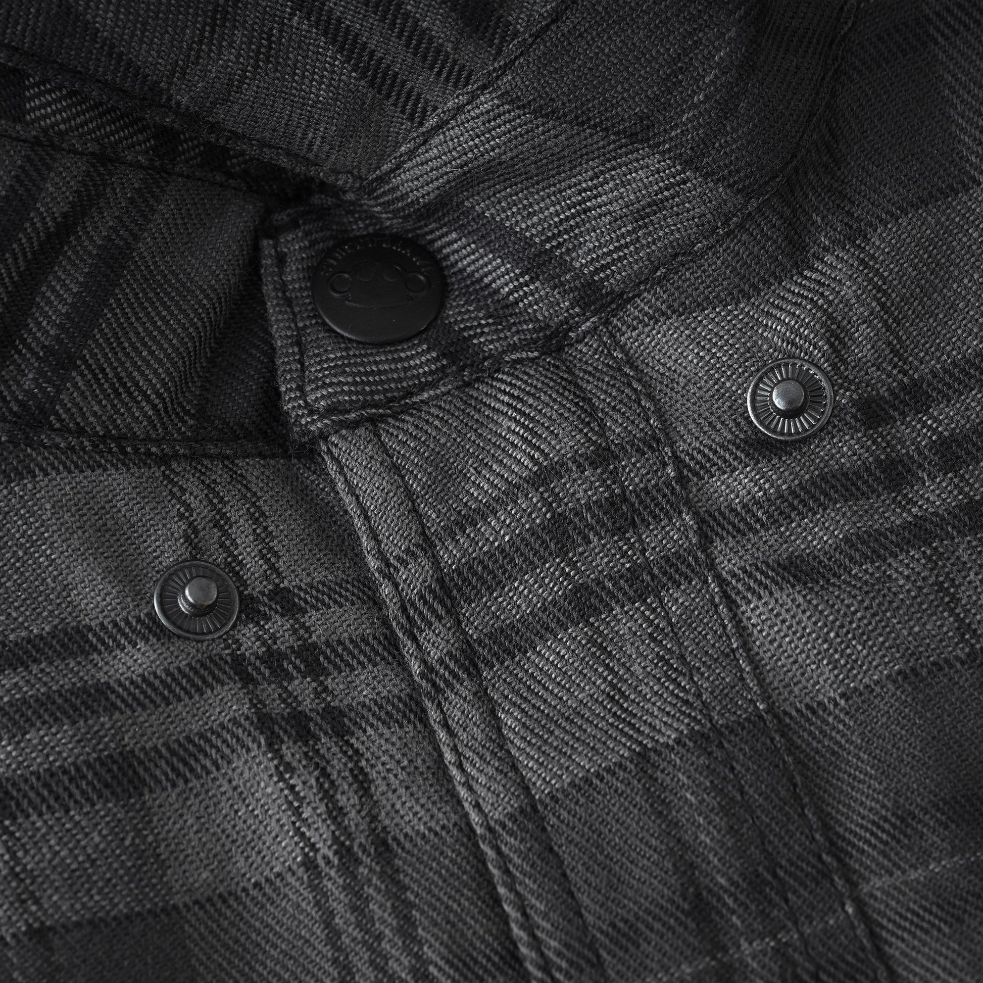 Close-up of a dark gray and black plaid fabric jacket or overshirt with black snap buttons, showing the detailed woven texture and stitching.