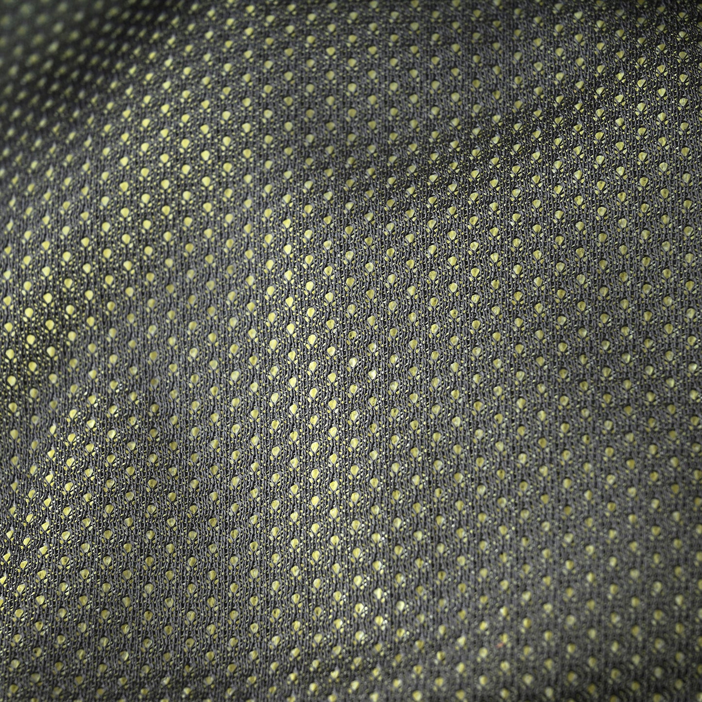 A close-up of a woven fabric with a repeating small geometric dot pattern in muted yellow-green on a dark charcoal gray background.