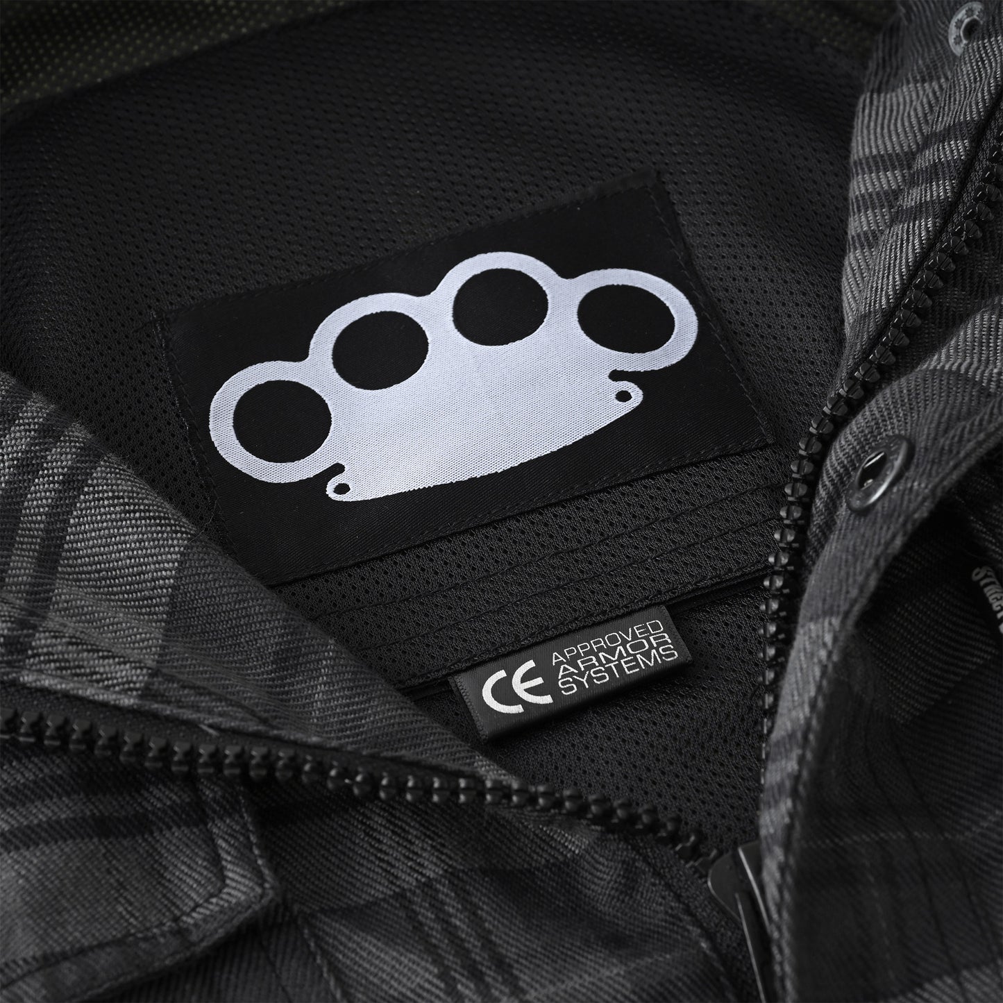 Close-up of a black zippered jacket interior showing a black patch with a white brass-knuckle style logo and a small black tag reading 'APRON HAZMAT SYSTEMS'.