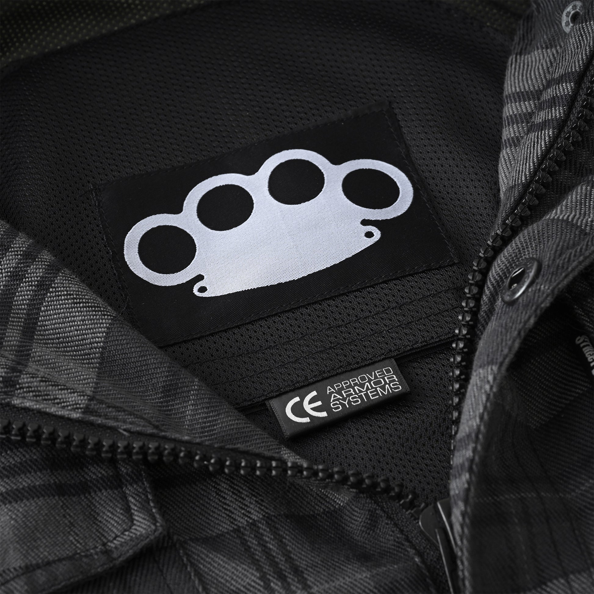 Close-up of a black zippered jacket interior showing a black patch with a white brass-knuckle style logo and a small black tag reading 'APRON HAZMAT SYSTEMS'.